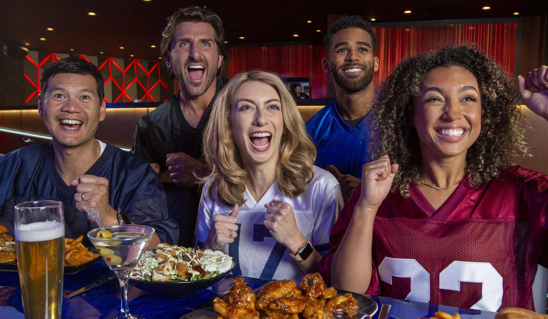 A group of five friends celebrating while watching a sports event. They are seated at a table with food including wings and salads. They are wearing football jerseys and showing excitement, with drinks on the table.