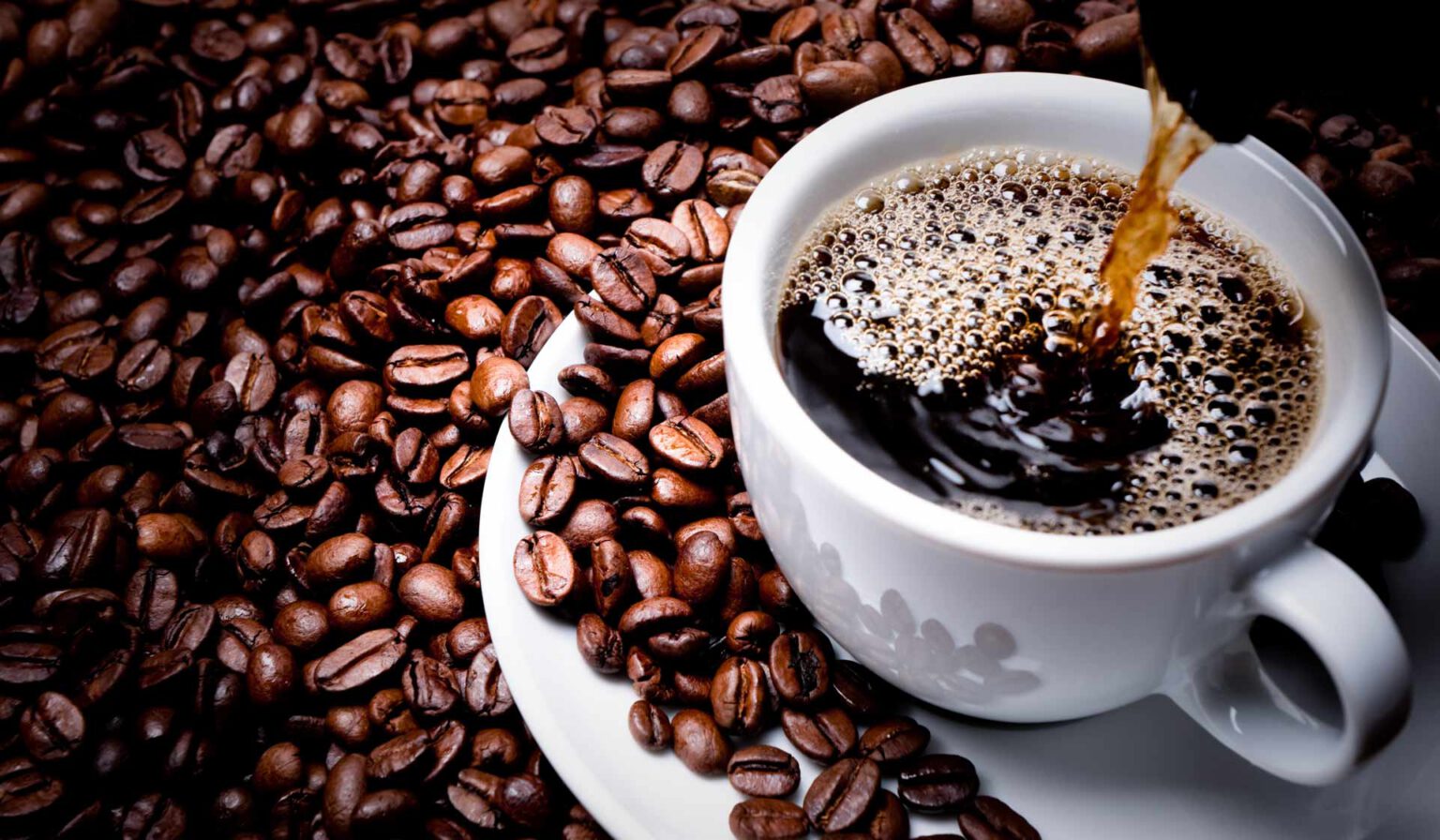 A cup of coffee being poured with many coffee beans in the background.