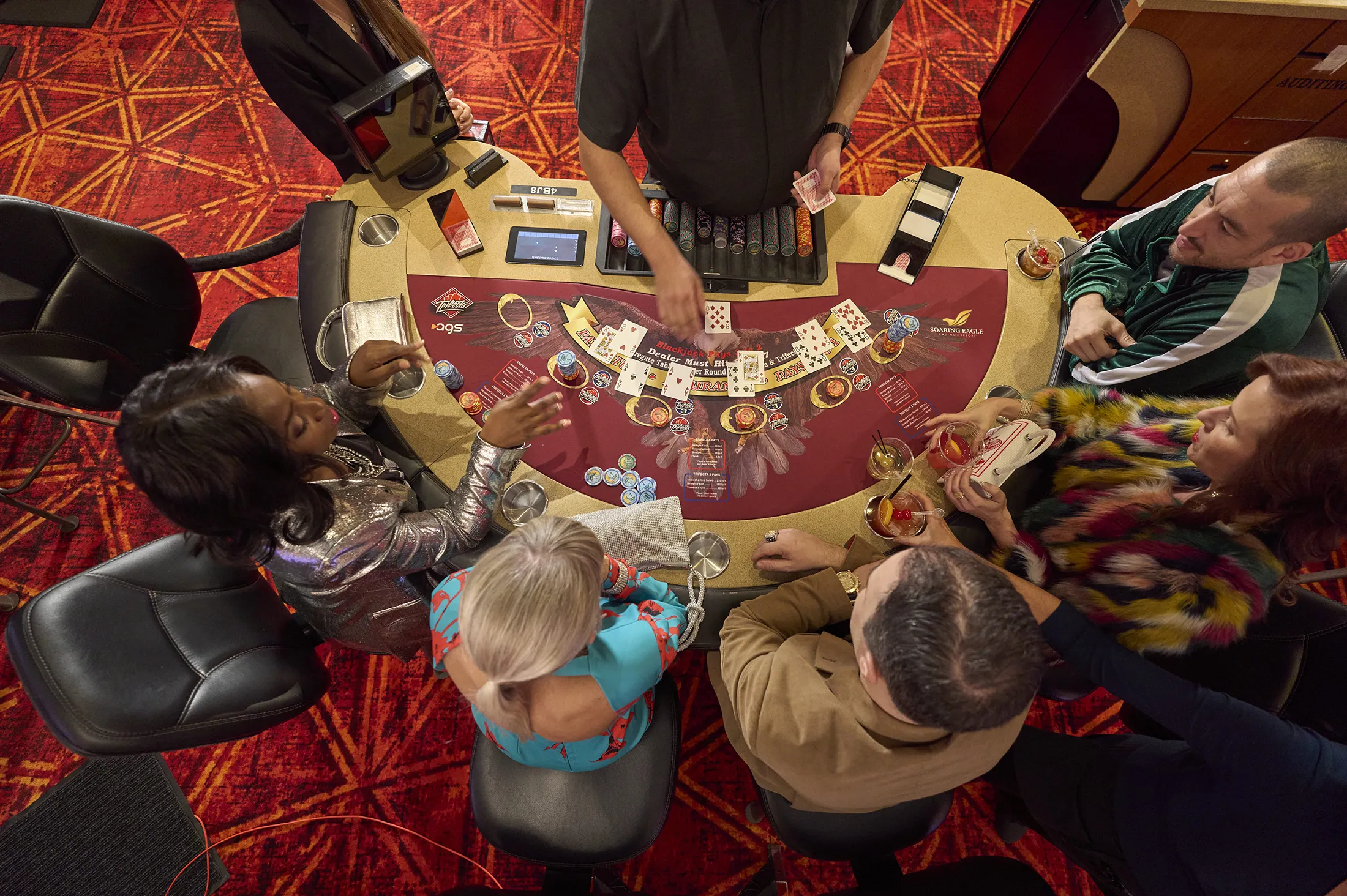 An overhead shot of men and woman playing poker at a table and a dealer laying down cards.