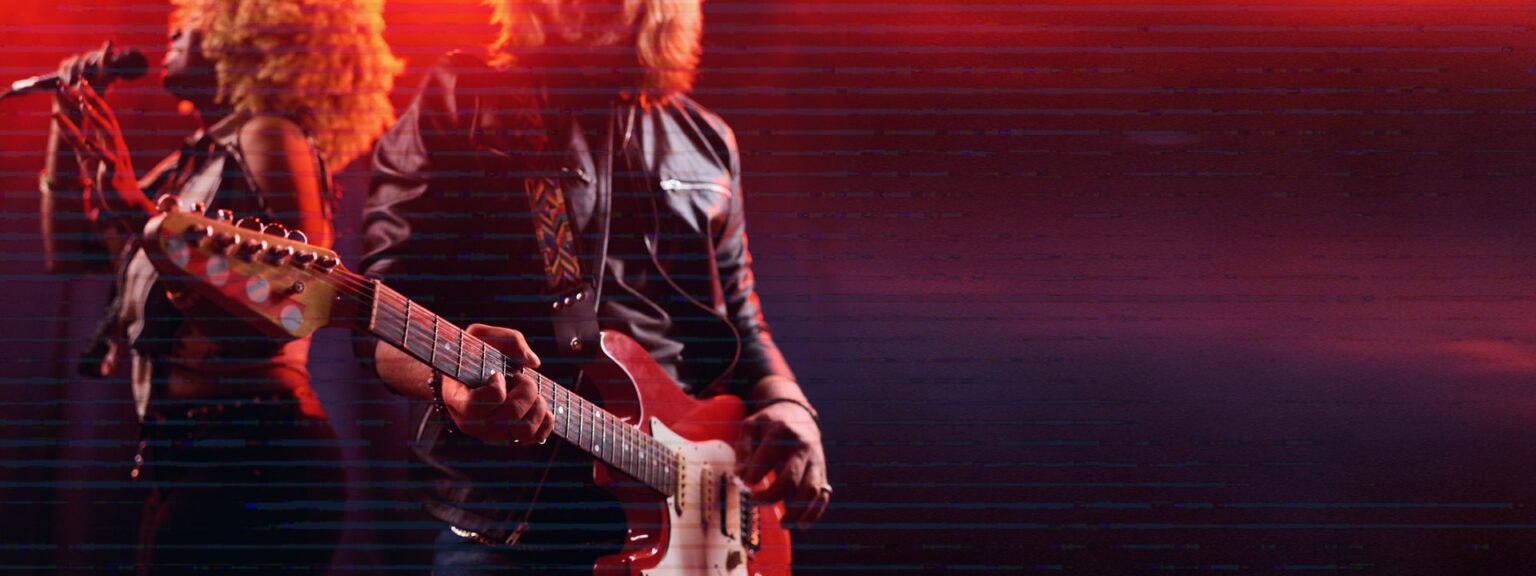 A guitarist playing an electric guitar on stage, with vibrant red lighting illuminating the scene. In the background, a singer with curly hair is holding a microphone, contributing to the live music atmosphere.