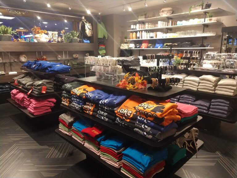 A retail display featuring neatly arranged stacks of colorful clothing, including t-shirts and hoodies, on multiple shelves. The setting has bright lighting and decorative elements, with a variety of other merchandise visible in the background.