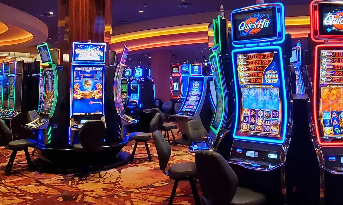 An interior view of a casino featuring multiple brightly lit slot machines. The machines display colorful graphics and are arranged in rows on a patterned carpet. The ambient lighting creates a vibrant atmosphere.