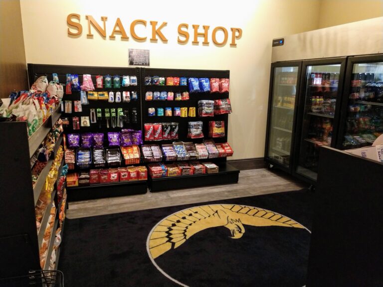 Interior of a snack shop featuring shelves filled with various snacks and drinks. The sign above reads "SNACK SHOP." A large rug with a logo is visible in the center of the floor, and glass refrigerators display cold beverages nearby.